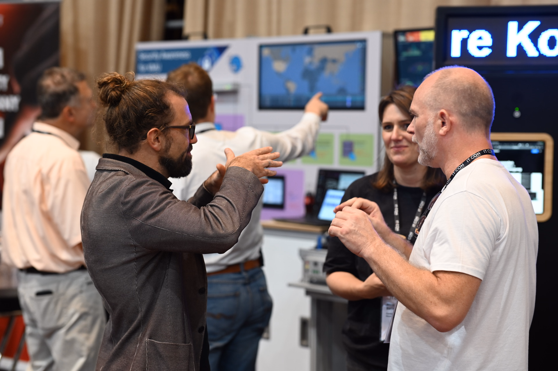 Three people exchange ideas at the exhibition booth, with digital applications visible on screens and tablets in the background.